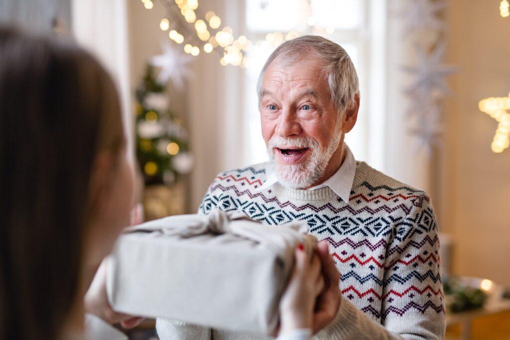 Bilde av overrasket mann (far, bror, mann, bestefar, forelder eller pappa) som får en gave av en ung jente. Huset i bakgrunnen er pyntet til jul med julelys og juletrær. Mannen som mottar gaven er glad.
