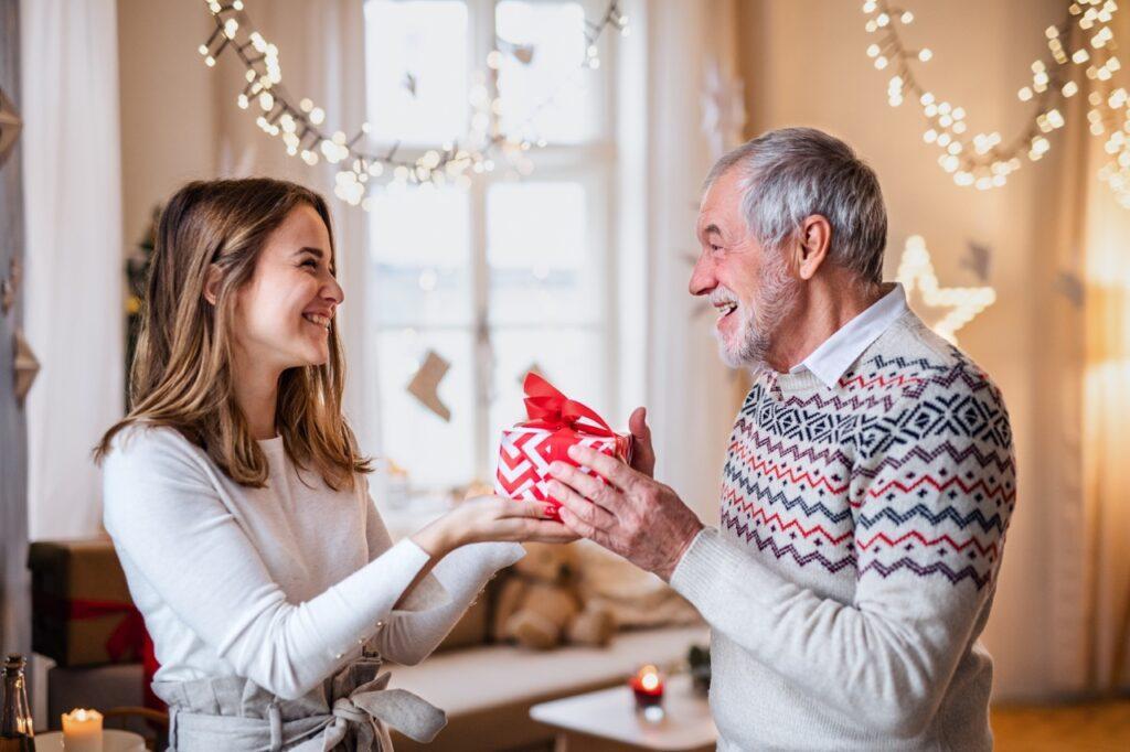En ung dame som smiler til en eldre mann som gir henne en julegave pakket i rødt og hvitt julepapir. Vi har samlet 50 julegavetips til hele familien slik at du enkelt kan finne julegave til henne, julegave til han, julegave til barn, julegave til par og billige julegaver til under 500 kroner.