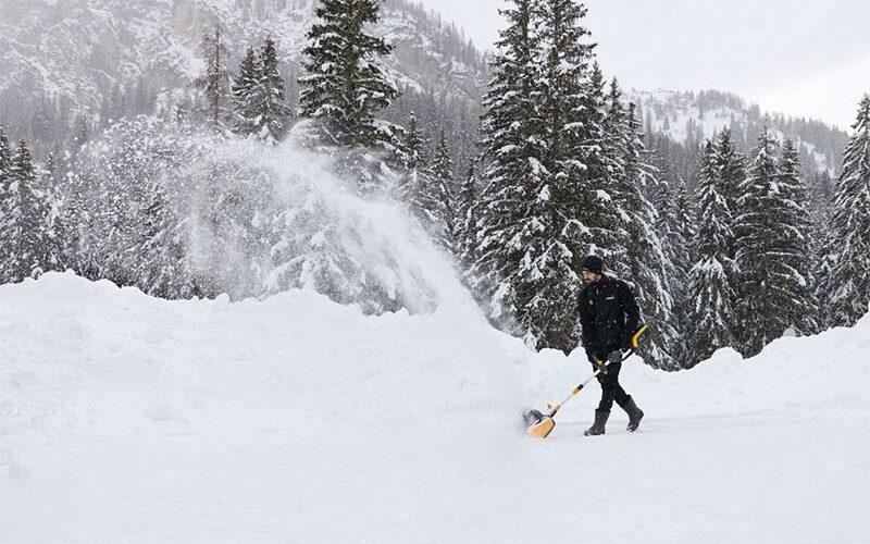 Bilde av mann i snødekket natur med fjell og trær som bruker en elektrisk snøskuff/ elektrisk snøfreser.