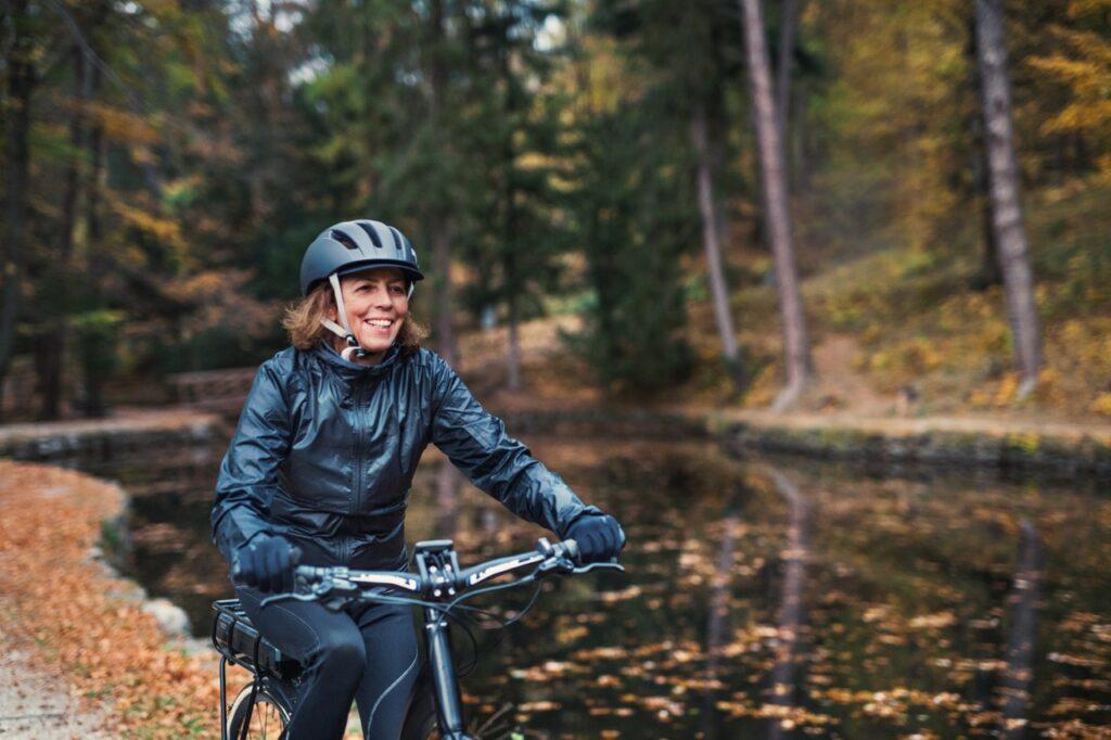Bilde av dame ute i naturen, ved et vann og med høstfarger og oransje blader. Damen sykler på en elsykkel langs et vann, smiler og har på seg sykkelhjelm og regnklær. Til saken vår om elsykkel hvor vi avslører elsykkel til dame som er best i test i 2022 og 2023.