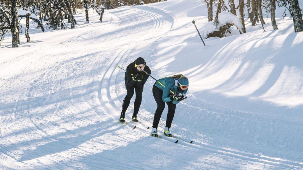 Bilde av to personer på vei ned en bakke på snø i vinterlandskap med felleski på beina.