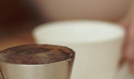 Bilde av verdens sunneste kaffe, gresk kaffe. En hand holder i håndtaket til en liten kanne med gresk kaffe på vei til å helle den i en hvit kopp på et bord.