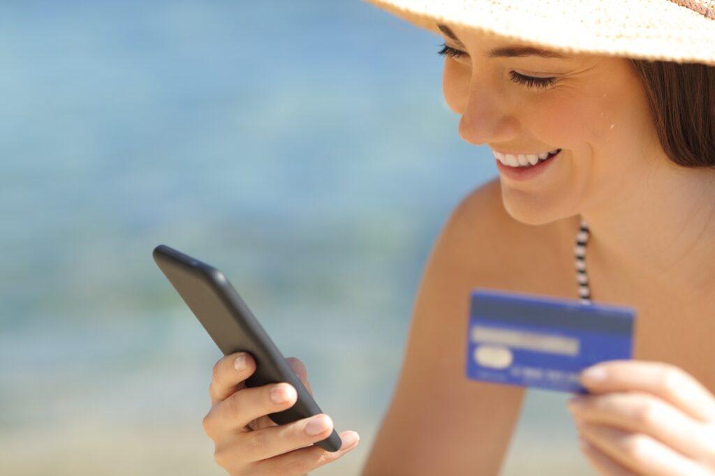 Nærbilde av smilende dame som holder en telefon i den ene hånden og et blått kredittkort i den andre hånden. Blått hav i bakgrunnen. Vi forteller deg fordelene med å bruke kredittkort når du skal ut å reise.