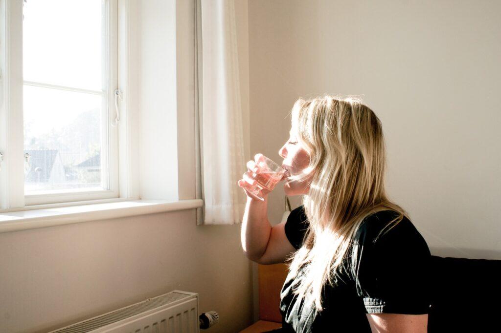Kvinne som sitter og drikker vann av gjennomsiktig glass foran vindu, med solen i ansiktet. Vi forteller hva vanntabben som kan sende deg opp i vekt er.