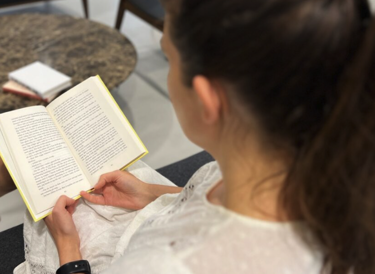 Brunette i hvit kjole sitter i en sofa hvor hun leser en bok. I bakgrunnen kan man se flere bøker ligge på et bord. Vi forteller om fem positive og overaskende helseffekter lesing har.