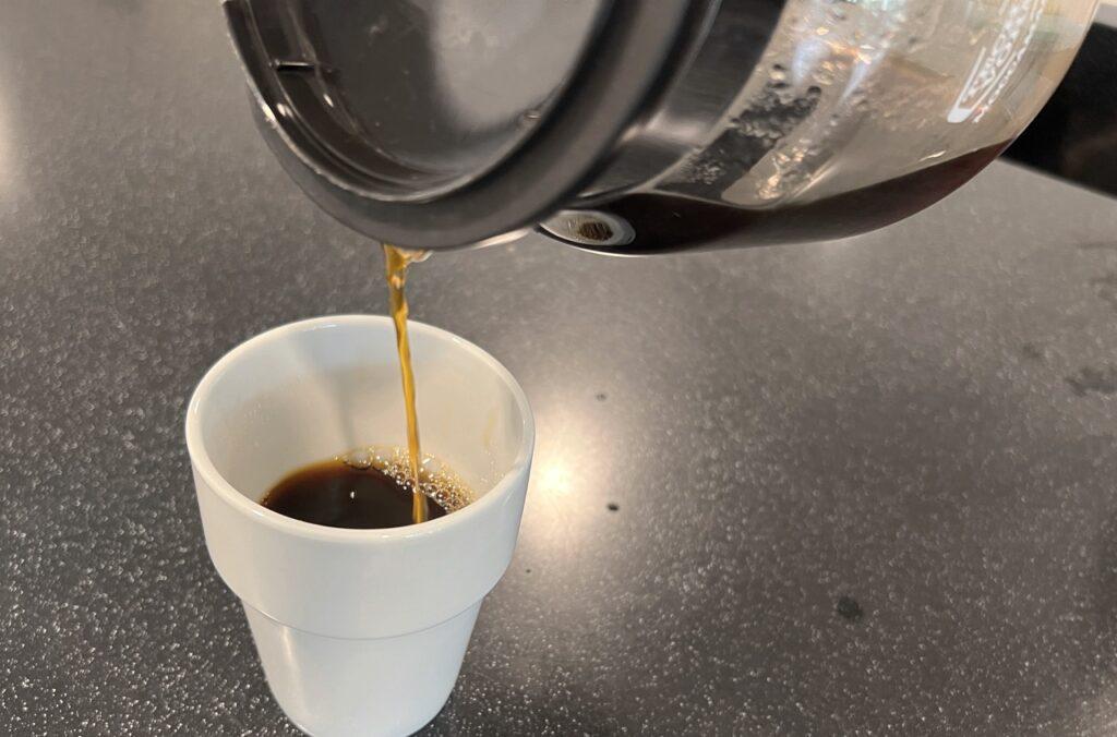 Bilde av en hvit kopp på en sort benk. I koppen er det litt kaffe og en kaffekanne heller i mer kaffe. Vi forteller hvorfor det er sunt å drikke kaffe.