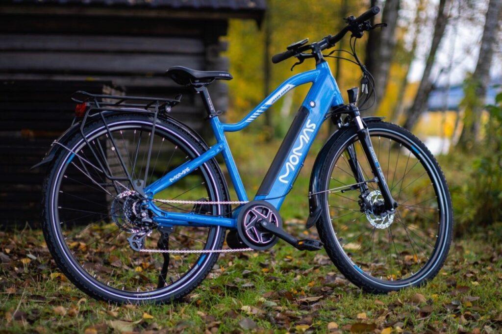 Bilde av momas-elsykkel på tilbud i 2023. Elsykkelen er blå og står ute i natur med høstblader.