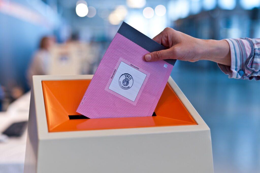 Valg 2023: Kommune- og fylkesvalget nærmer seg. På bildet ser du en orange stemmeboks hvor noen putter en rosa stemmeseddel i. Vi har funnet svar på vanlige spørsmål angående stemming slik at du føler deg forberedt før du går til valglokalene uavhengig av om du skal stemme i Oslo, Bergen, et lite tettsted, hjemmefra eller fra utlandet.