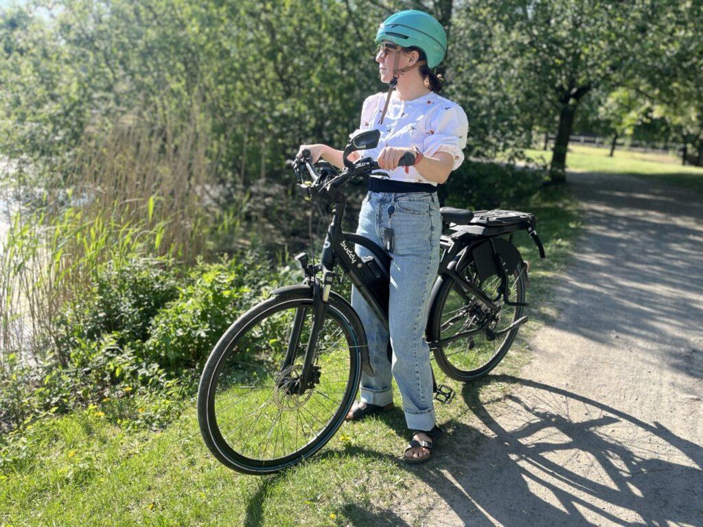 Bilde av dame som står med en elsykkel til dame fra Buddy Bike. Hun har på seg en hvit bluse, dongeribukser og en blå sykkelhjelm og står med elsykkelen sin på en gressplen med et vann i bakgrunnen i sol og sommervær. Vi forklarer hva som kjennetegner en sykkel og elsykkel til dame!