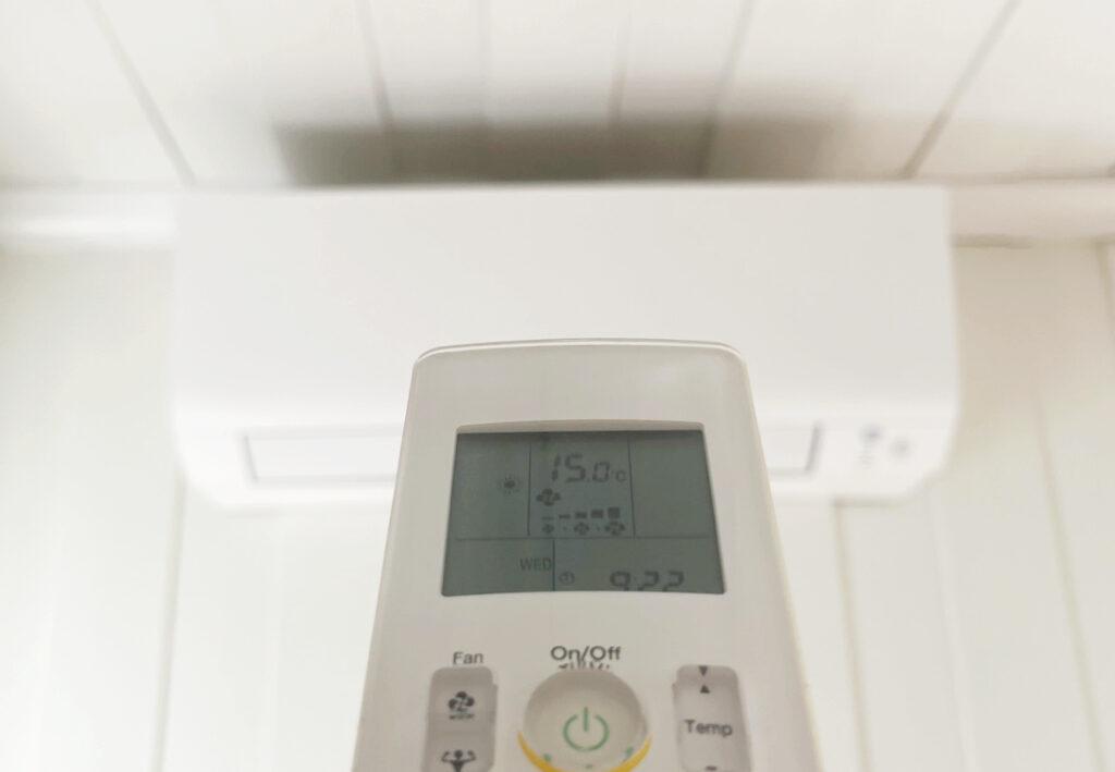 Nærbilde av fjernkontrollen til en varmepumpe med en luft til luft-varmepumpe (blurry) i bakgrunn. Vi forteller hvor mye en luft til luft-varmepumpe bruker, og hvor mye su kan spare på å installere en varmepumpe hjemme,