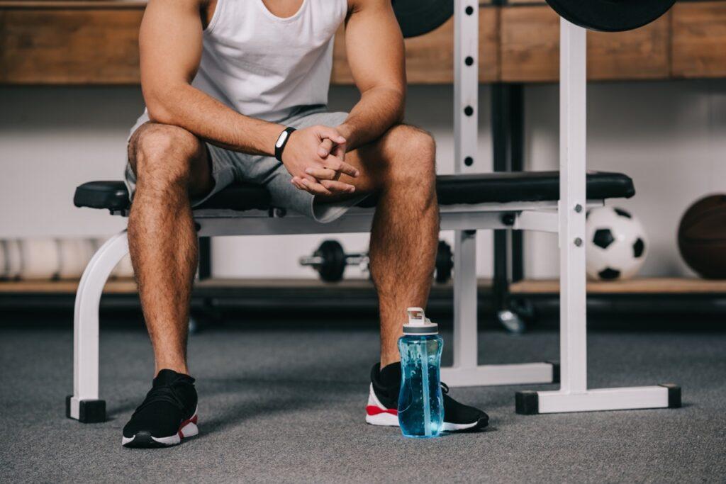 Person som sitter på en treningsbenk som er del av et multigym. Bilde er kuttet slik at man ikke ser ansiktet til personen. På gulvet ved siden av føttene står en blå vannflaske. Vi har funnet multigym-apparatene som er best i test. Velg blant Finnlo Bio Force, Abilica Multi 4ST og Abilica Multi 970.