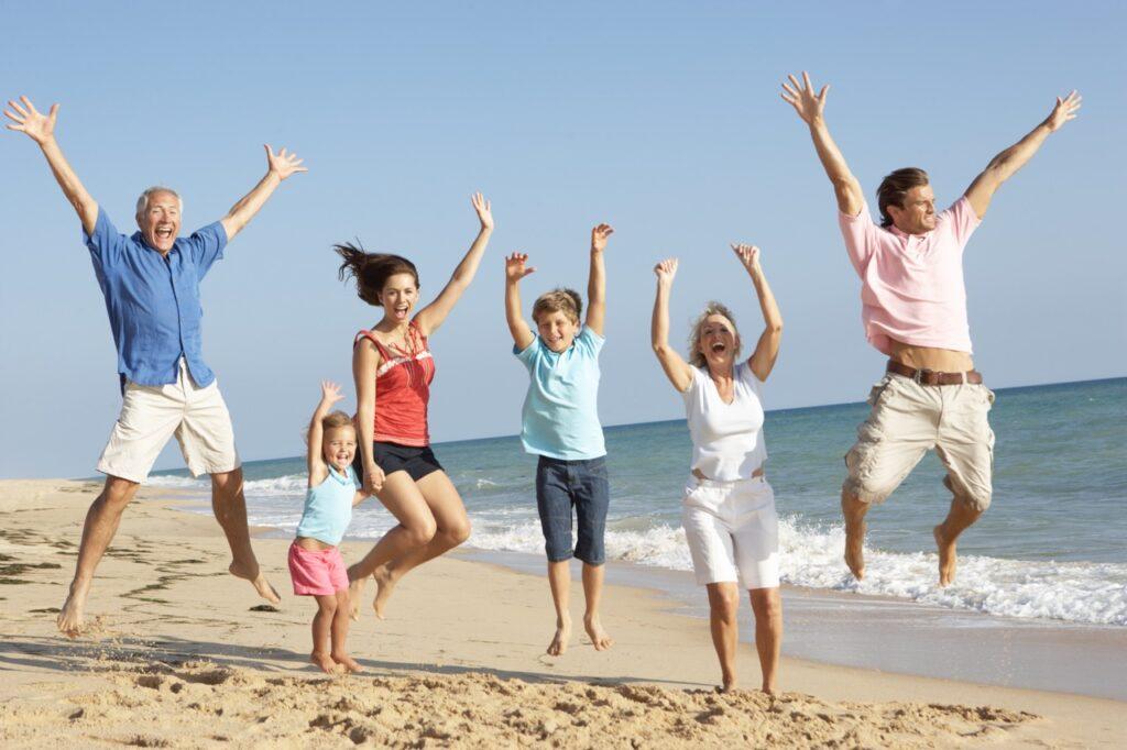 En flergenerasjonsfamilie nyter en dag på stranden, med alle som hopper gledelig i luften. Gruppen inkluderer en eldre mann i en blå skjorte og hvite shorts, en kvinne i en rød tanktopp og svarte shorts, en ung jente i en lyseblå tanktopp og rosa shorts, en gutt i en lyseblå skjorte og mørke shorts, en eldre kvinne i en hvit skjorte og hvite shorts, og en mann i en rosa skjorte og beige shorts. I bakgrunnen ser vi en klar blå himmel og havbølgene som forsiktig treffer den sandete stranden. Vi forklarer hvilke fordeler du får ved å bestille pakkereiser.