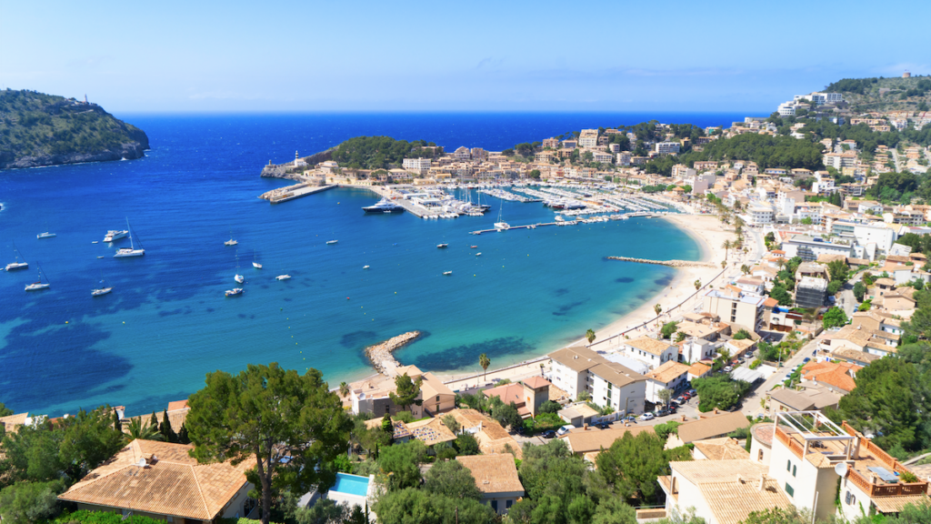 Bilde av en liten middelhavsby på den spanske øya Mallorca. På bilde ser du knallblått hav, en hvit strand. båter på havet og en koselig liten by som strekker seg opp i fjellet. Vi har samlet restplasser til mallorca slik at du kan finne billig reise til syden.