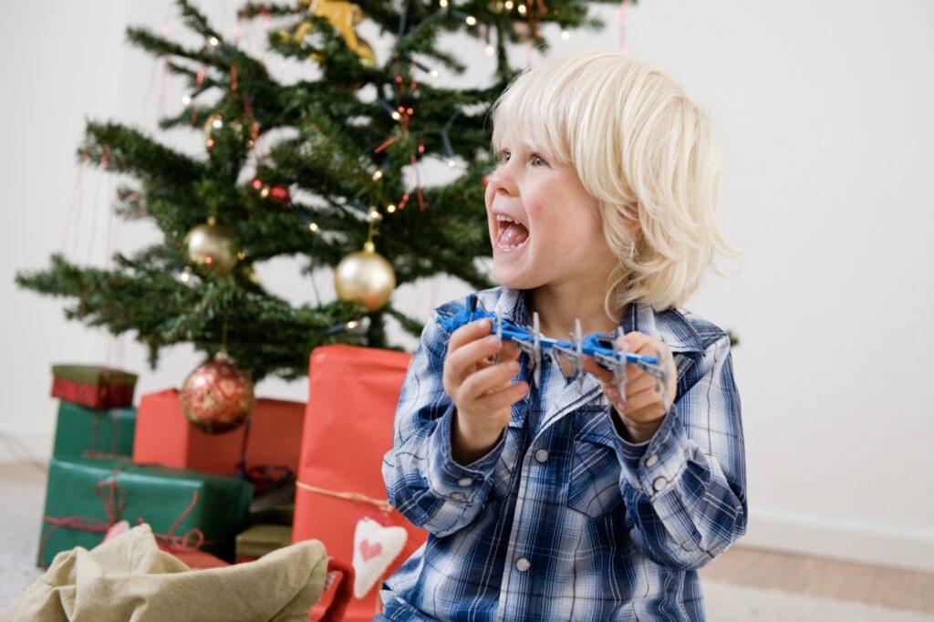 Bilde av smilende gutt med en julegave i hendene. Gutten sitter i en stue foran et juletre med julegaver under, i flere forskjellige farger.
