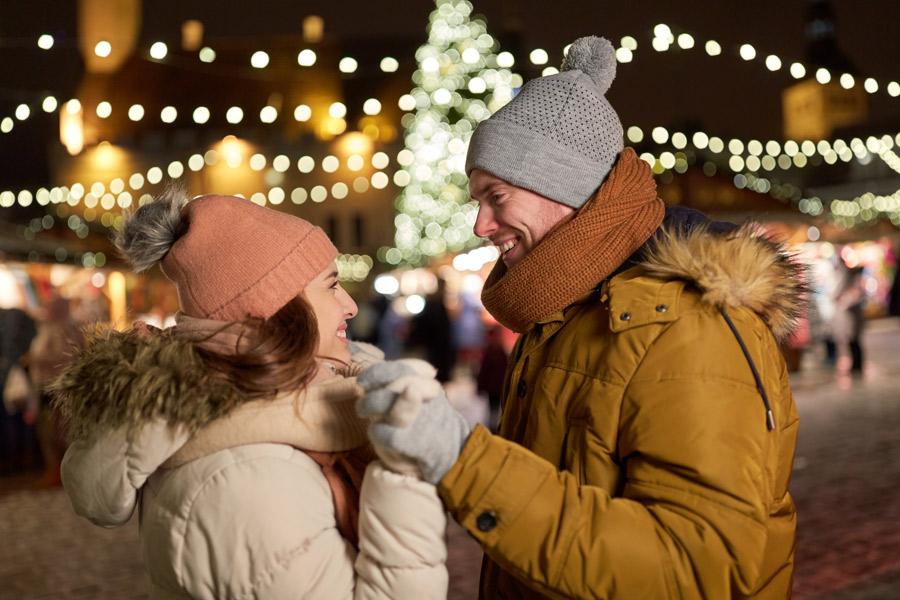 Mann og dame som står mot hverandre og ser hverandre i øynene mens de holder hender. De har på seg vinterklær og i bakgrunnen kan man se julelys fra et julemarked i Tallin. Vi har samlet billige pakkereiser til Europas mange julemarked i Tallin, Gøteborg, Berlin, Wien, Praha, London, Kiel, København, Krakow og Gdansk.