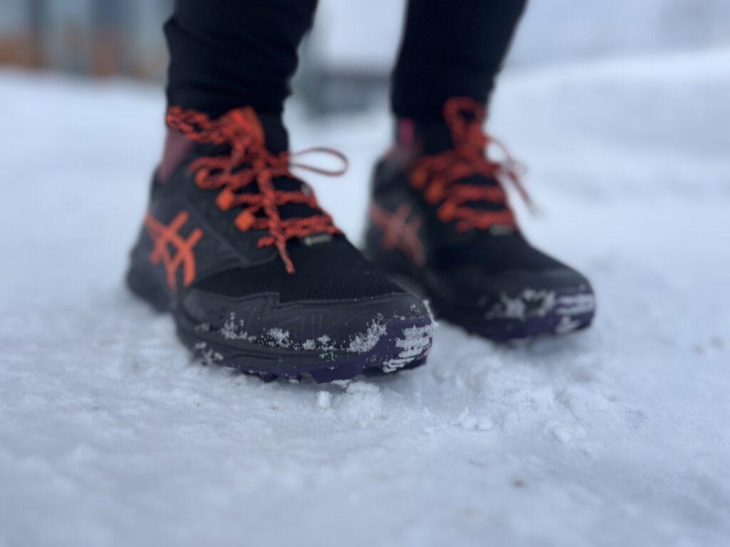 Bilde av person med asics-piggsko på føttene. Personen står med de svarte skoene med oransje lisser og detaljer ute i snø i et nabolag. Vo forteller hvor du kan finne billige piggsko på tilbud!