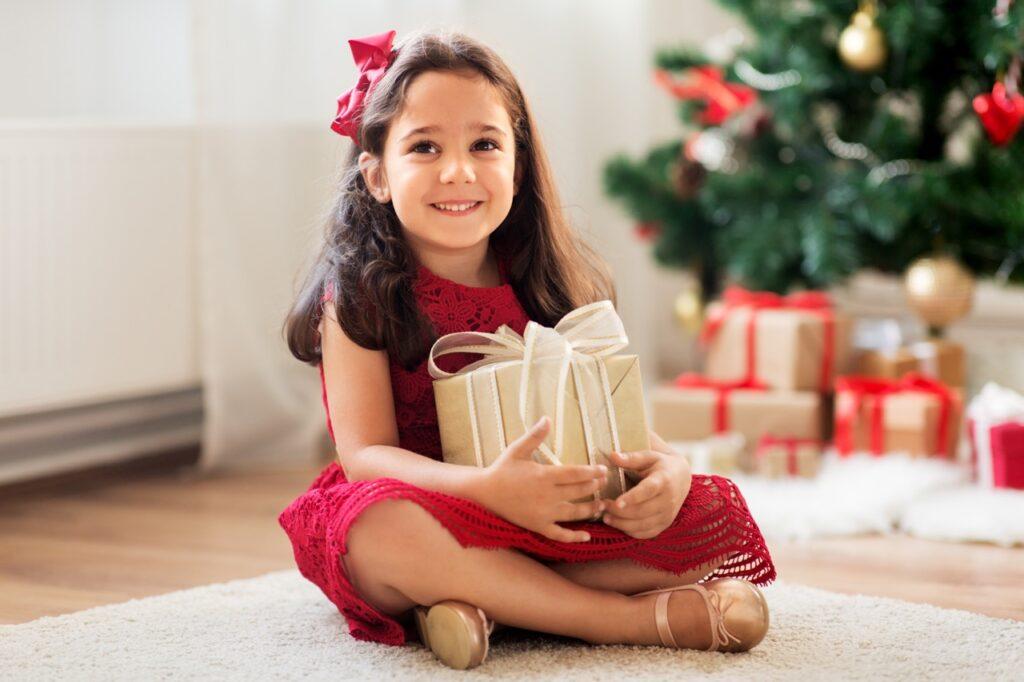 Bilde av liten jente i rød kjole, pyntet til julaften. Hun sitter på et lyst teppe med et pyntet juletre i bakgrunnen, og under juletreet ligger det mange innpakkede julegaver. Vi gir deg tips til julegaver til barn: Jente 3-5 år. Både billige og dyrere!
