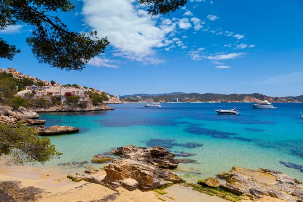 Bilde av krystallklart hav på Mallorca i Spania, rundt havet ser du en gyllen strand som er omringet av kliper og grønne busker. Vi hjelper deg med å finne restplasser til Mallorca slik at du kan finne billig reise til syden. Vi gir deg også fem tips til hva du kan se og gjøre på øya, samt svarer på ofte stilte spørsmål om Mallorca.