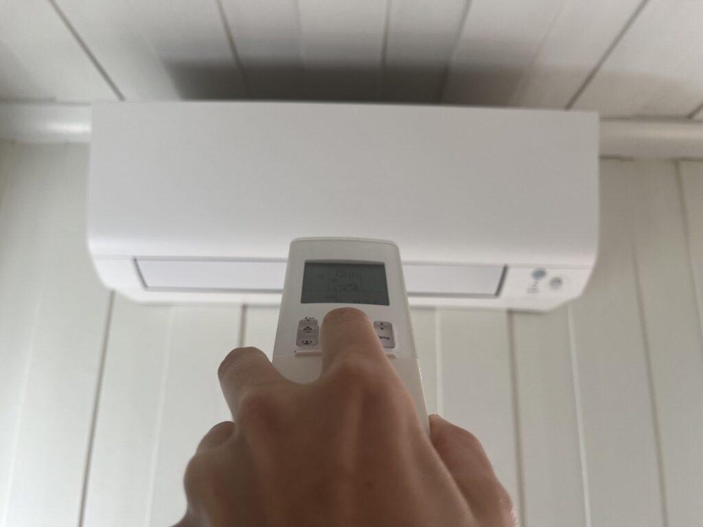 Bilde av en hand som trykker på en knapp på en varmepumpe-fjernkontroll og som sikter den mot en luft til luft-varmepumpe som henger oppe på en vegg. Vi har snakket med eksperten som forteller hvilken innstilling du bør bruke for å bruke varmepumpen mest effektivt, samt hvilken innstilling du ikke må bruke på varmepumpen om vinteren. Vi firteller også hvor mye sytrøm en varmepumpe bruker i forhold til annen opp varming, hvor mye strøm du sparer med en varmepumpe samt hvor mye strøm du sparer med en varmepumpe om vinteren.