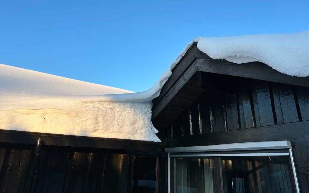 Bilde av snø på tak på et svart hus, med blå himmel i bakgrunnen. Vi forteller hvilke regler som gjelder når det kommer til snø på tak, samt når det er ditt ansvar å fjerne den.