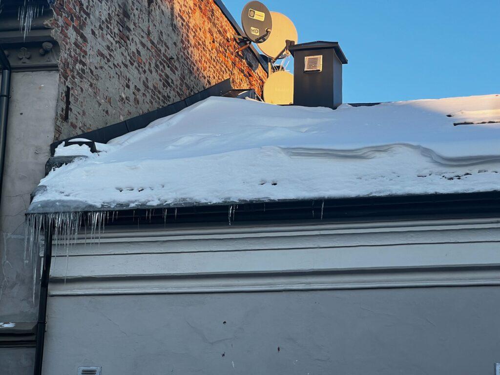 Bilde av snø og is på et hustak. Det er et hvitt murbygg med en svart takrenne og med blå, skyfri himmel i bakgrunnen. Vi forteller hvilke regler som gjelder når det kommer til snø på tak, samt når det er ditt ansvar å fjerne den.