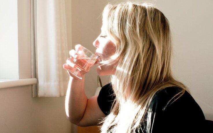 Kvinne som sitter og drikker vann av gjennomsiktig glass foran vindu, med solen i ansiktet. Vi forteller hva vanntabben som kan sende deg opp i vekt er.