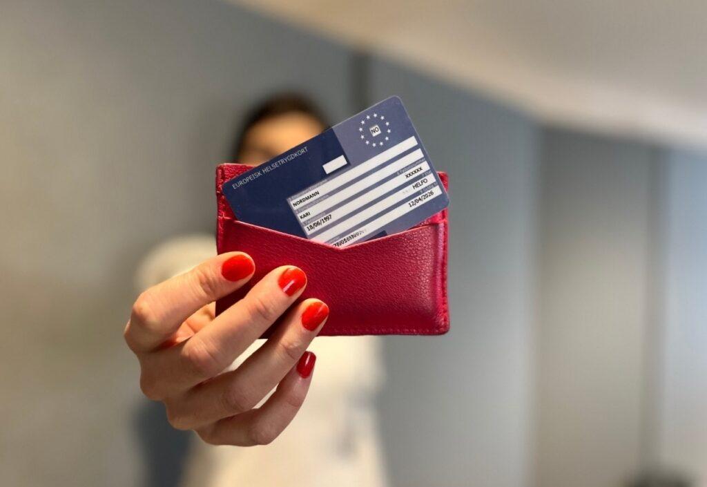 Person med rød neglelakk som holder opp en rosa lommebok med et Europeisk helsetrygdkort i. Vi har snakket med eksperten for å kunne fortelle deg alt du må vite om Europeisk helsetrygdkort.