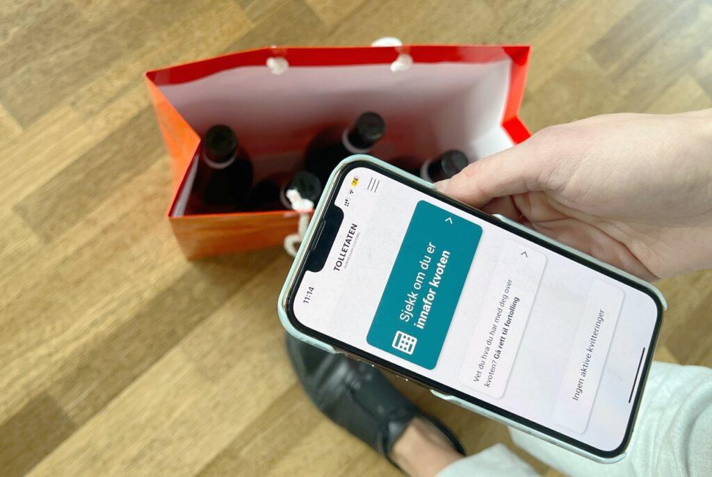 Person som bruker kvote-appen til Tolletaten for å se om innkjøpet av vin er innenfor kvoten. På gulvet står en pose med tre vinflasker. Vi har snakket med Tolletaten som forklarer kvote-reglene i Norge på blant annet alkohol, tobakk, og verdigrensen du må følge.