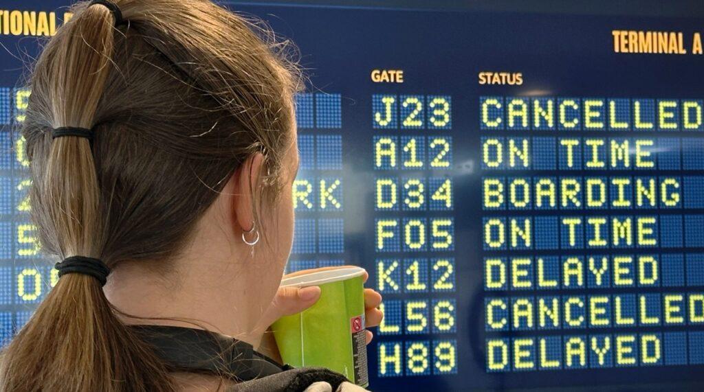 Bilde av en person med brunt hår som holder en grønn pappkopp men hen ser på tavla over fly på en flyplass. Flere fly er forsinket. Matkupong er noe man kan få av flyselskapet, men dette burde man si nei til. Vi forklarer.