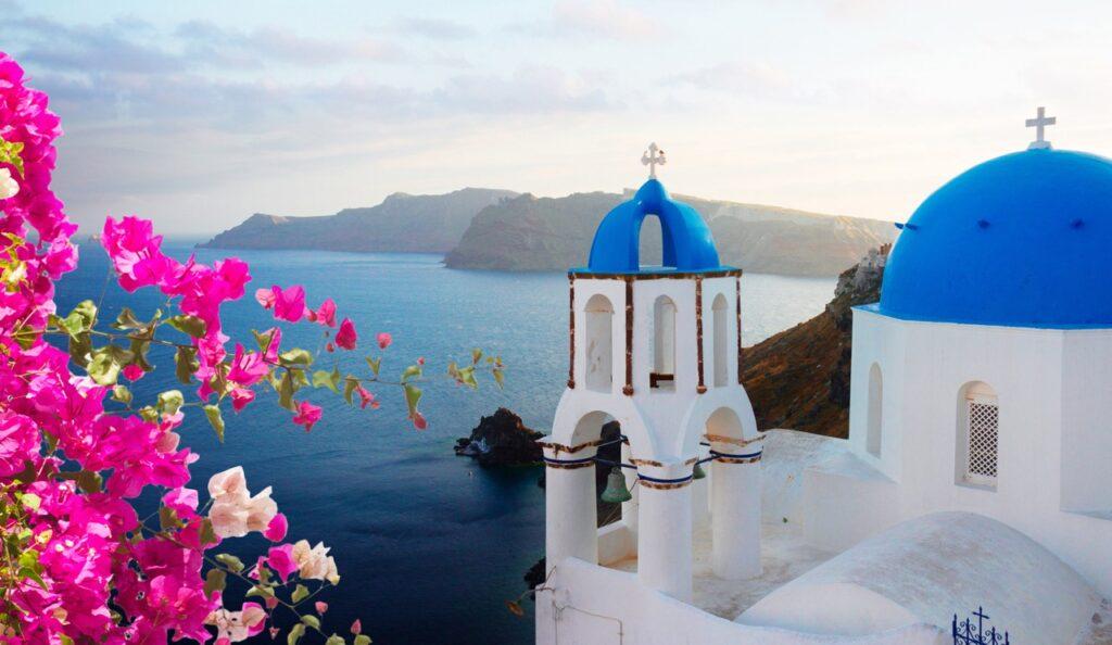 Bilde fra Santorini med utsikt over Egeerhavet. På høyre siden ser du et klassisk hvitt murbygg med blå kuppel på, mens på venstre side er rosa blomster som står i kontrast til de blå takene. Vi har skrevet om fem ting man bør se og gjøre på Santorini i Hellas, samt avslører hvilket hotell på Santorini som er kåret til det beste. Her finner du også restplasser til Santorini og billige reiser til de greske øya