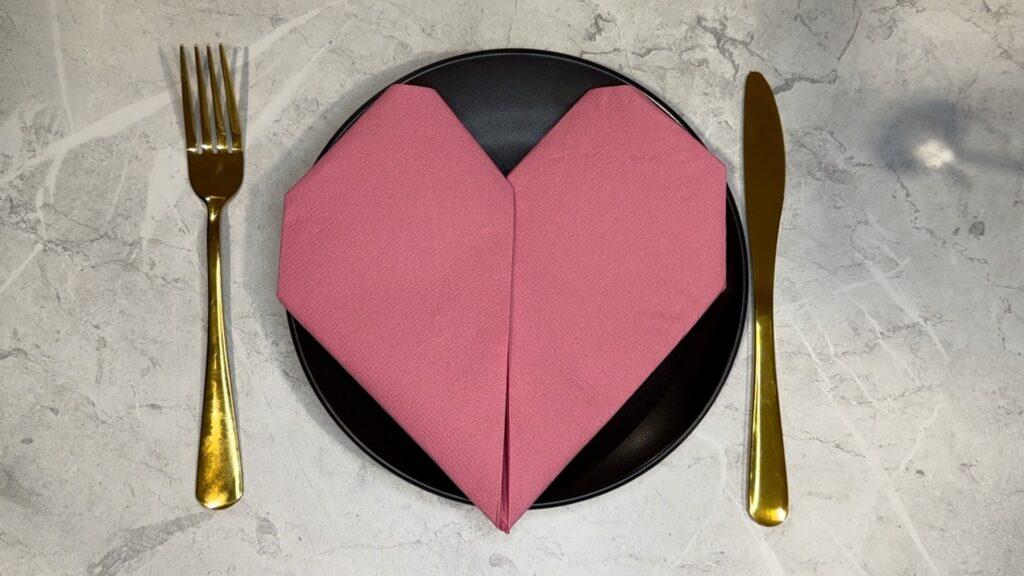 En rosa serviett brettet som et hjerte på en tallerken med bestikk ved siden av.