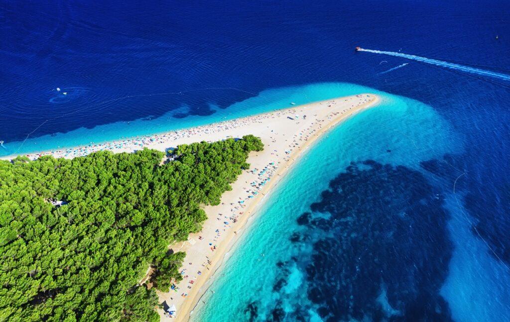 Bilde av Zlatni Rat-stranden som ligger på øya Brac ved byen Bol i Kroatia. Stranden er spesiell og kan ligne et gyllent horn som er omringet av krystallklart hav. Mot land kan man se grønne trær. Vi forklarer hva du må se og gjøre på Brac, som ligger utenfor Split i Kroatia, samt hjlper deg med å finne billige reiser til Brac, hvorav restplasser er populære.