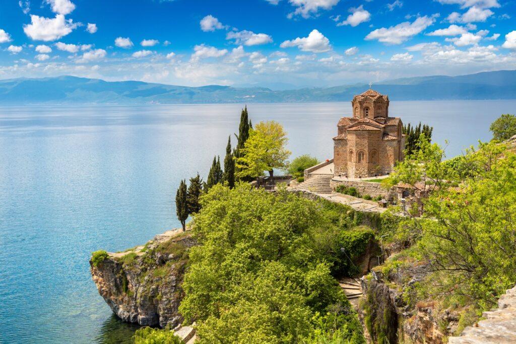 Et vakkert bilde av kirken St. John at Kaneo som står på en klippe over Ohridsjøen i Nord-Makedonia. Kirken, med sin tradisjonelle bysantinske arkitektur, dominerer bildet med sine detaljerte mursteinsmønstre og et fremtredende kuppeltak. Den ligger omgitt av frodige grønne trær og busker, med trappetrinn som leder ned mot vannet. I bakgrunnen strekker det store, rolig blå vannet seg mot fjellene under en delvis skyet himmel. Nord-Makedonia har blitt rangert som landet i Europa som er billigst for nordmenn å feriere i. Vi har samlet fem ting du bør se og gjøre i landet, samt hjelper deg med å finne billige reiser ved hjelp av restplasser.