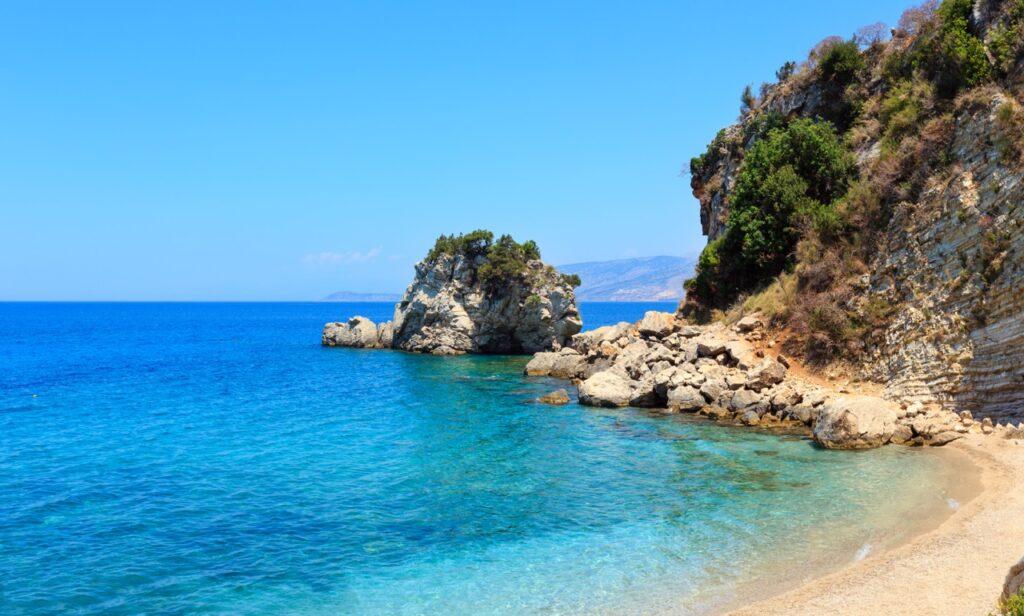 Bildet viser en vakker kystlinje med klart blått vann, en steinete strand og frodige grøntområder på klippene i den populære byen Saranda i Albania. Midt i vannet står en stor stein med busker og trær som vokser på toppen. I bakgrunnen kan man se fjell og en klar, blå himmel uten skyer. Vi har samlet restplasser til Albania slik at du kan finne billige reiser til landet. Vi svarer på spørsmål som; Er Albania trygt? og, hva koster en øl i Albania? Vi forteller også om populære destinasjoner i Albania som Saranda, Durres, Vlorë og Tirana.