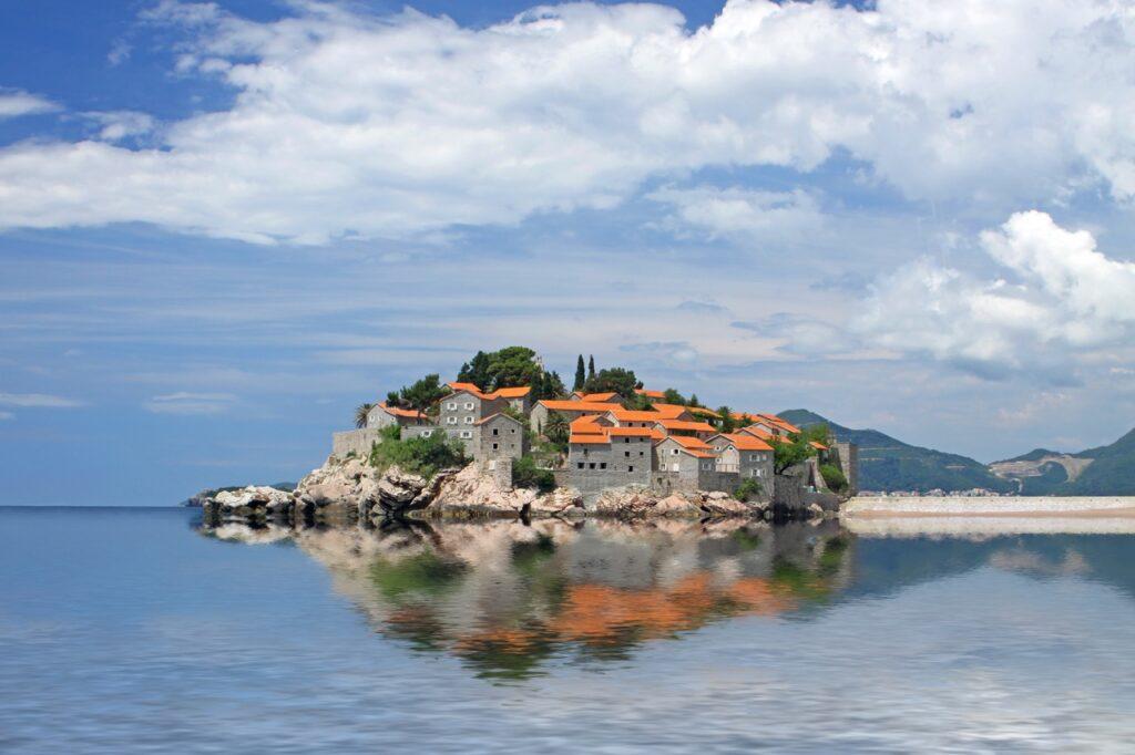 Panoramautsikt over øya Sveti Stefan i Montenegro, med sine karakteristiske oransje taktegl på gamle steinbygninger. Øya er omgitt av klipper og reflekteres vakkert i det rolige havet. Over øya er en skyet himmel, som gir en dramatisk bakgrunn. Montenegro er et spennende land langs Middelhavet som er rangert til å være et billig land for nordmenn å feriere i. Vi hjelper deg med å finne pakkereiser som inkluderer fly og hotell, samt restplasser til Montenegro.