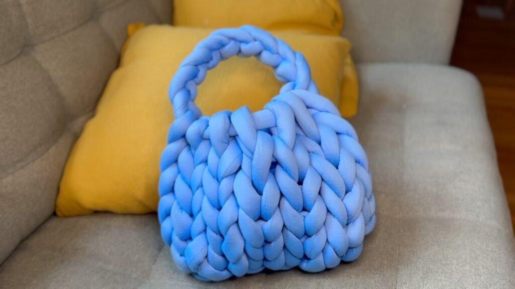 En blå veske strikket i chunky garn ligger i en grå sofa med en gul pute bak. Vi har oppskriften på hvordan man strikker vesken med hendene.