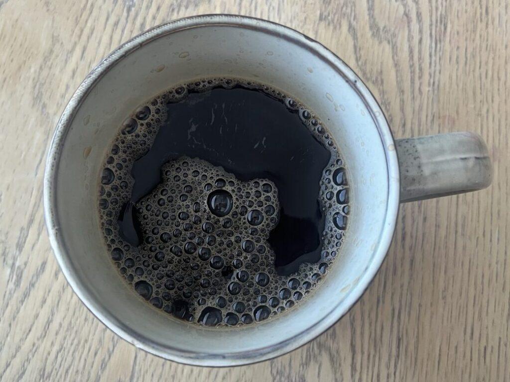 Bilde av en beige keramikk-kopp med sort kaffe på spisebord i tre. Vi forteller om overraskende forskning som tilsier at det ikke er lurt å drikke kaffe etter lunsj og hvorfor! Vi forklarer også hvor mye lenge koffeinen i kaffe blir værende i systemet og hvor lenge du bør vente etter din siste kopp kaffe før du legger deg til å sove om kvelden fordi den har innvirkning på søvnen din!