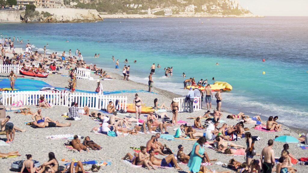 Bildet viser en travel strand full av mennesker som soler seg og bader i havet. Strandgjestene ligger på fargerike håndklær og bruker parasoller for å beskytte seg mot solen. Noen personer er i vannet og svømmer, mens andre sitter i oppblåsbare båter nær vannkanten. Flere bygninger og trær kan sees i bakgrunnen. Vannet har en klar turkis farge, og himmelen er klar uten skyer, noe som indikerer en varm og solrik dag. Hetebølger herjer i Europa, spesielt langs Middelhavet. Land som Hellas, Kypros og Tyrkia opplever ekstremvarme med temperaturer opp mot 40 grader. Vi gir deg alternative destinasjoner hvor du er mindre utsatt for ekstreme temperaturer. Reis til Sopot i Polen, Nord-Makedonia, Algarvekysten, Kanariøyene eller på norgesferie i år.