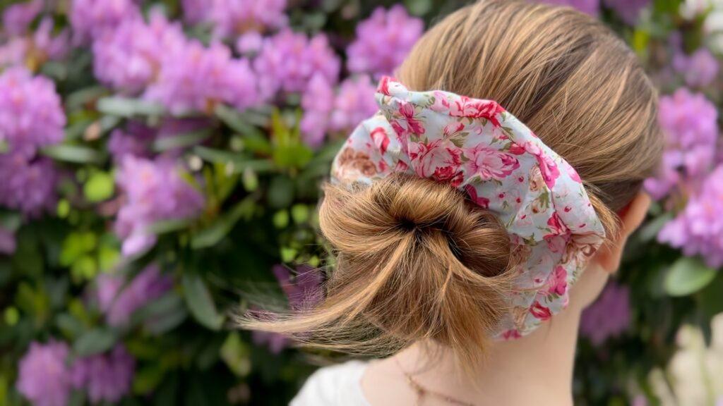 En jente med mørk blondt hår satt opp i en topp med en scrunchie i blomstrete stoff. Hun står foran en rhododendronbusk.