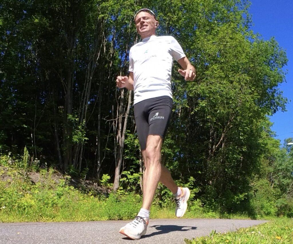 Bildet viser en mann som løper utendørs på en solrik dag. Han har på seg en hvit t-skjorte med logoer og svarte shorts. På det venstre bildet ser vi mannen smilende og løpende mens han tar en selfie. Det høyre bildet viser mannen bakfra mens han løper på en asfaltert vei omgitt av grønn natur og trær. Dette bildet formidler glede ved løping og en aktiv livsstil utendørs i vakre omgivelser. Løpeeksperten Andreas Gossner fra løpetrening.no svarer, Hvordan lager du ditt eget løpeprogram?