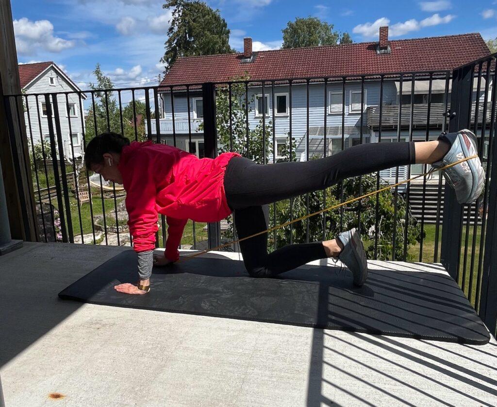 En kvinne (Cecilie Aasland) trener bein på en treningsmatte utendørso. Hun har på seg en rosa treningsjakke, svarte tights, løpesko fra Hoka i lyseblå, og utfører en styrkeøvelse med ett ben plassert påtreningsmatten, mens hun skyver den andre bak seg med en strikk som motstand. I bakgrunnen er det grå rekkehus, hager, trær, og blå himmel. Studioet har et industrielt utseende med treningsutstyr og ringer hengende fra taket. Løpeeksperten forklarer hvorfor må man trene styrke når man er glad løping?