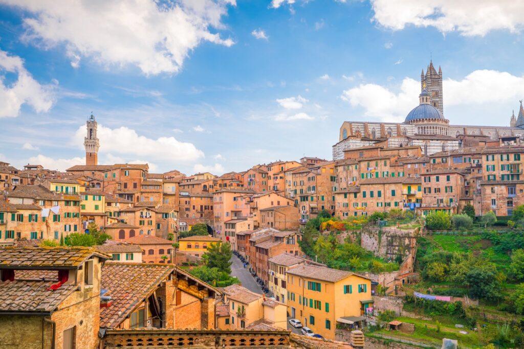 Bilde av den pittoreske byen Siena, Toscana i Italia på en solrik dag med blå himmel og noen skyer. Byen er bygget på en ås, og de historiske bygningene er tett sammen med karakteristiske røde tak og varme jordfarger. Øverst i bildet kan man se den imponerende katedralen i Siena med sin kuppel og gotiske detaljer. Flere grønne områder og hager kan ses mellom bygningene, noe som gir byen et frodig utseende. Vi har laget en omfattende guide til Toscana i Italia hvor vi blant annet forteller om fem populære byer som Firenze, Pisa, Siena, Montepulciano og Volterra. Vi gir deg også forslag til tre kjøreruter du kan ta for å oppleve vingårder og resten av Toscana. La deg inspirere av den italienske regionen som fikk sitt eget TV-program; «Den store matfesten - Toscana»