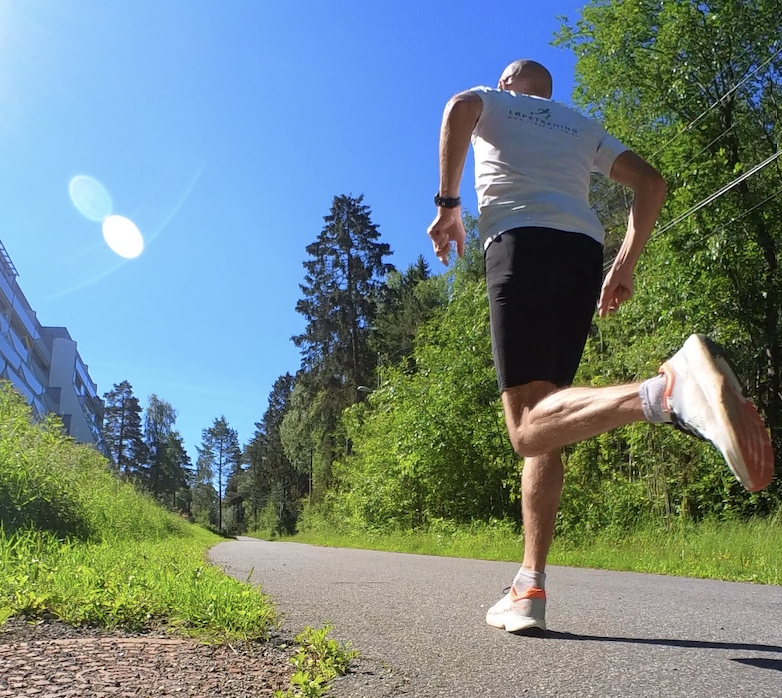 Bildet viser en mann som løper utendørs på en solrik dag. Han har på seg en hvit t-skjorte med logoer og svarte shorts. På det venstre bildet ser vi mannen smilende og løpende mens han tar en selfie. Det høyre bildet viser mannen bakfra mens han løper på en asfaltert vei omgitt av grønn natur og trær. Dette bildet formidler glede ved løping og en aktiv livsstil utendørs i vakre omgivelser. Løpeeksperten Andreas Gossner fra løpetrening.no svarer, Hvordan blir du en robust løper?