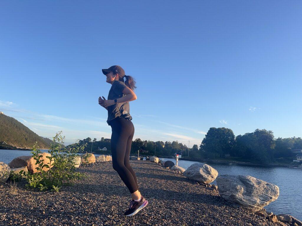 Bilde av en dame/kvinne som løper/ jogger ute på en pir som leder ut i Oslofjorden i naturen. I bakgrunnen ser man grusvein hun løper på, store steiner som omkranser piren, en øy, sjø og grønne, frodige trær. Kvinnen smiler og har på seg en NY Yankees-cpas, en svart tights, og en grå t-skjorte. Skoene er svarte med farge-innslag av lilla, hvitt, rød, og oransje og er løpesko som er best i test fra Brooks. Personen trener seg opp til å løpe 1 mil (10 kilometer) ved hjelp av et treningsprogram med både vanlig løping og intervaller. Løpeekspert Andreas Gossner forteller hvordan du kan legge opp et treningsprogram for å trene deg opp til å løpe 10 kilometer enten du er erfaren eller nybegynner!