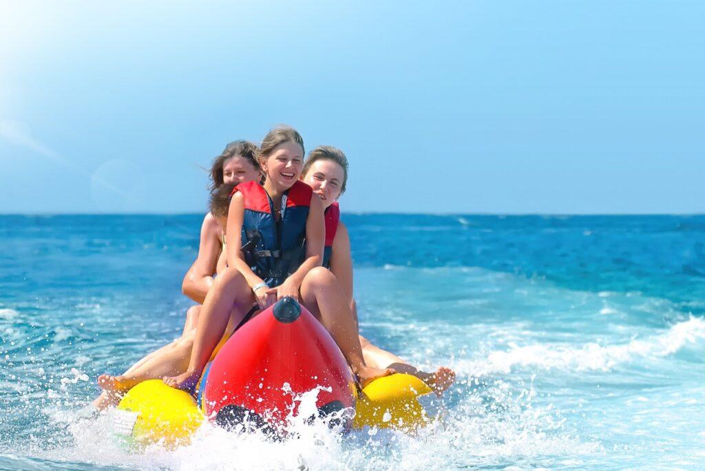 Bildet viser tre personer som sitter på en oppblåsbar bananbåt og ler mens de blir dratt gjennom vannet. De har på seg redningsvester, og vannet spruter opp rundt dem mens de nyter den spennende vannaktiviteten. Havet rundt dem er klart og blått, og himmelen er lys og skyfri. Her får du ti ting du kan gjøre på Gran Canaria, Kanariøyene