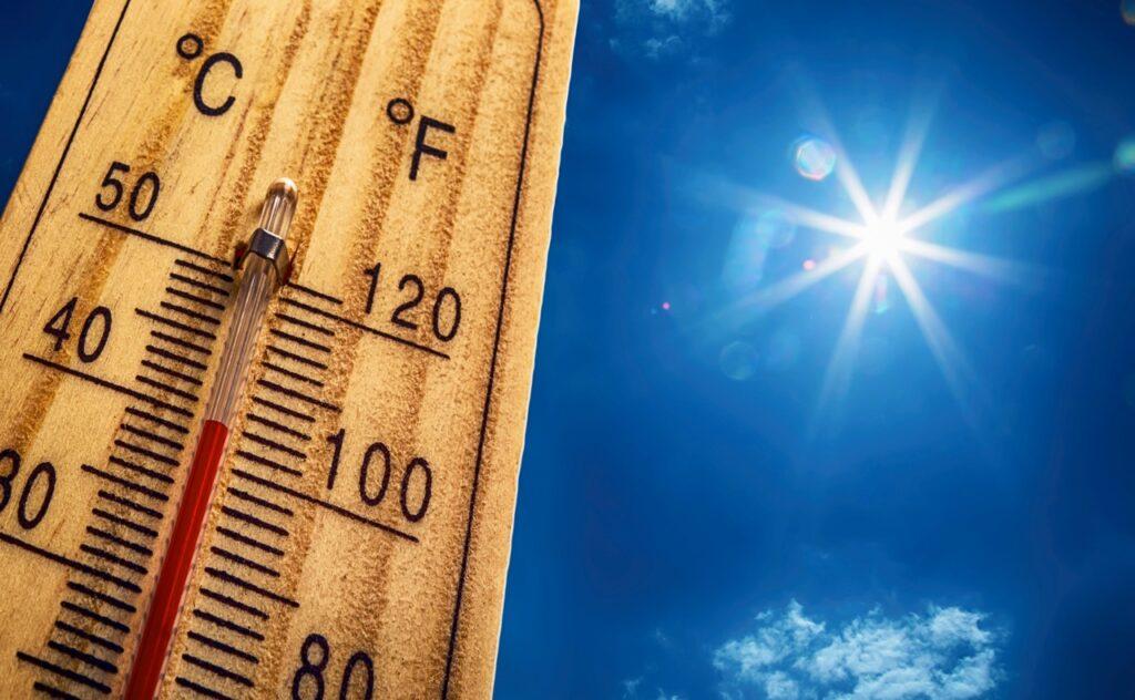Et nærbilde av et termometer som viser høye temperaturer, med en klar blå himmel og strålende sol i bakgrunnen. Her får du tips til hvordan du forebygger heteslag, også kalt solstikk, i ekstremvarmen. Vi forklarer symptomene på solstikk og hvem som er spesielt utsatt.