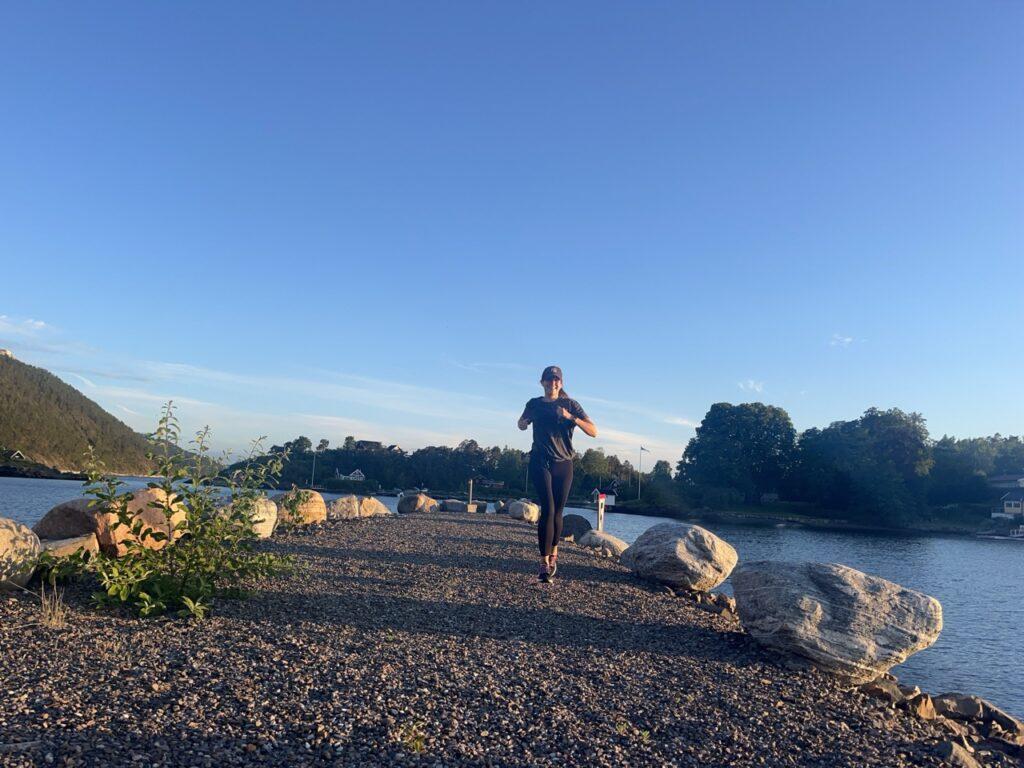 Bilde av en dame/kvinne som løper/ jogger ute på en molo som leder ut i Oslofjorden i naturen. I bakgrunnen ser man grusveien hun løper på, store steiner som omkranser moloen, en øy, sjø og grønne, frodige trær. Kvinnen smiler og har på seg en NY Yankees-caps, en svart treningstights, og en grå t-skjorte. Skoene er svarte med farge-innslag av lilla, hvitt, rød, og oransje og er løpesko som er best i test fra Brooks. Løpeekspert Andreas Gossner fra Løpetrening.no forteller at det å løpe langtur er noe av det som gir best fremgang på løpingen! Han forteller også hvordan det gjøres og hvorfor det gir god effekt på både kondisjon og fettforbrenning.