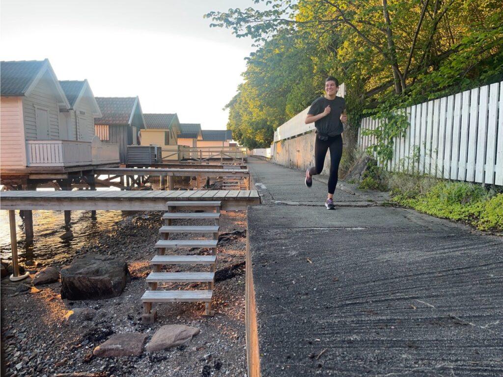 Bilde av en kvinne som jogger ved små badehus ved Oslofjorden. Hun har på seg en grå treningst-skjorte og svarte tights. Hun smiler bredt mens hun løper, og har en smartklokke på håndleddet. Bakgrunnen viser små badehus og blå himmel ved Oslofjorden. Solen skinner, og det er en lys og fin dag. Løpeekspert Andreas Gossner fra Løpetrening.no forteller hvordan du kan løpe raskt på lav puls!