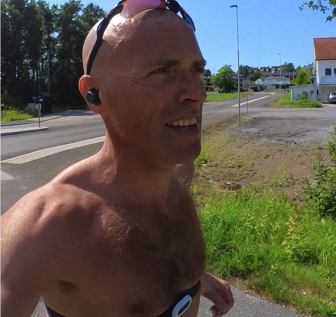 Bildet viser løpeeksperten Andreas Gossner fra løpetrening.no som løper utendørs på en solrik dag. Han har på seg en pulsmåler over bryster og solbriller fra Oakley til trening. På bildet ser vi mannen smilende og løpende på en asfaltert vei omgitt av grønn natur og trær mens han tar en selfie. Dette bildet formidler glede ved løping og en aktiv livsstil utendørs i vakre omgivelser. Løpetreneren svarer, Hvordan setter du ny personlig rekord på 10 km? Han gir deg også et treningsprogram som sikrer at du setter ny rekord på 10 kilometer!