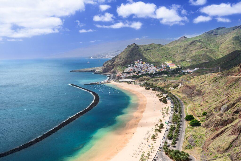 En vakker sandstrand på Tenerife, Kanariøyene, med gylden sand og turkisblått vann. Stranden er beskyttet av en lang molo som strekker seg ut i havet, og en rekke fargerike bygninger klatrer oppover den grønne åssiden i bakgrunnen. Flere små båter flyter på vannet nær moloen. Tenerife er den største Kanariøyen og har Spanias høyeste fjell, Teide. Hovedstaden er Santa Cruz de Tenerife. Her kan du finne billige reiser til Tenerife, samt hotell på den populære kanariøyen.